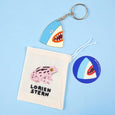 Keychain with a shark head in light blue with a red mouth and teeth, outlined in gold shown with a Lorien Stern bag and sticker