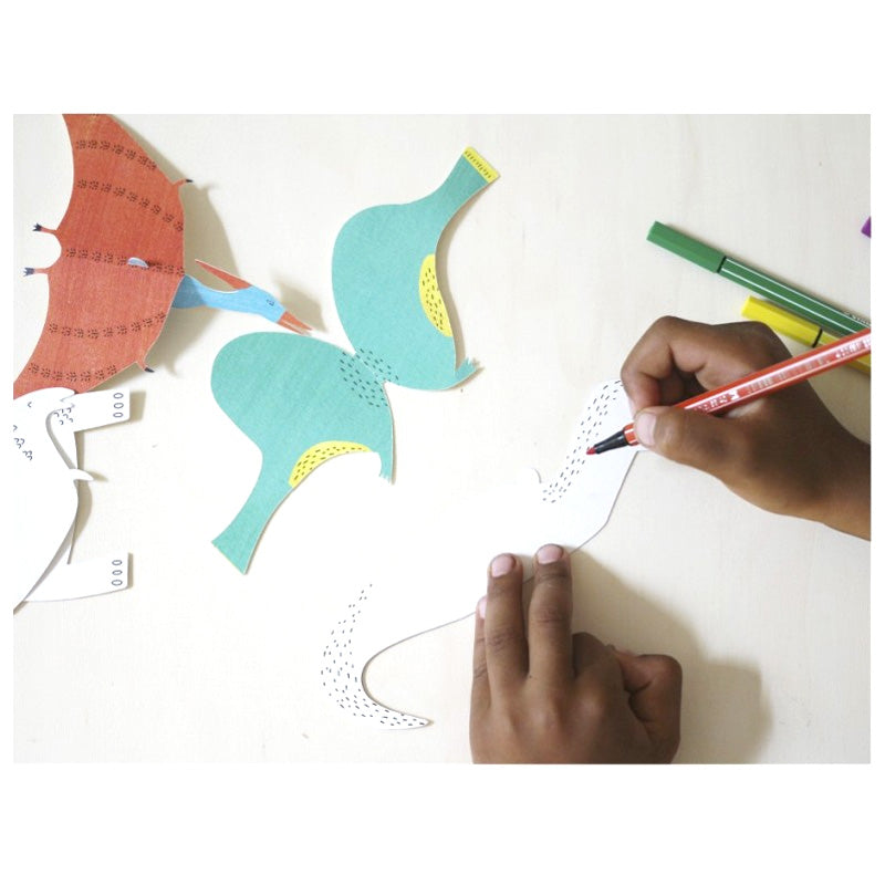 Paper dinosaurs craft kit being assembled with a child's hand coloring in the dinosaur