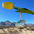 Gold keychain with a large gold toad with black spots shown hanging from a flower with a desert landscape in the background