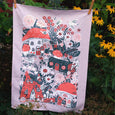 Tea towel with drawings by Phoebe Wahl showing tree stump and mushroom houses surrounded by flowers and gnomes hanging laundry and sweeping.