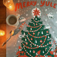 Phoebe wahl Merry Yule tea towel on the floor with a mug and spoon on top and candles around the sides