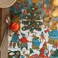 Tea towel with drawings by Phoebe Wahl of animals and people dancing and singing around a holiday tree, shown on a table with spoons and an apple on top