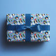 Present with a blue bow showing wrapping paper of penguins in winter hats holding presents and candy canes on a bright blue background.