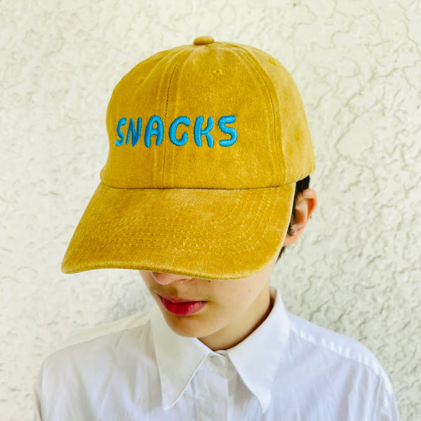 Baseball cap in ochre color with the word snacks in large turquoise embroidered text across the front