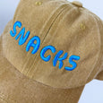 Baseball cap in ochre color with the word snacks in large turquoise embroidered text across the front