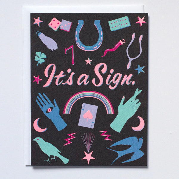 Greeting Card with black paper and with the words "It's a sign" in pink neon letters and illustrations of luck symbols around it in pastel colors