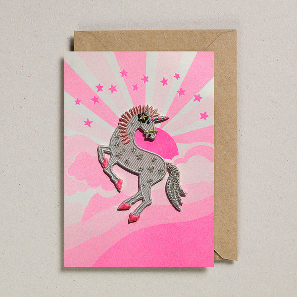 A unicorn iron on patch in white and pink attached to a paper greeting card showing a pink sunset