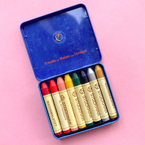 Eight stockmar wax stick crayons in a tin case with the colours: gold, silver, pearl pink, magenta, flame red, mid yellow, leaf green, indigo
